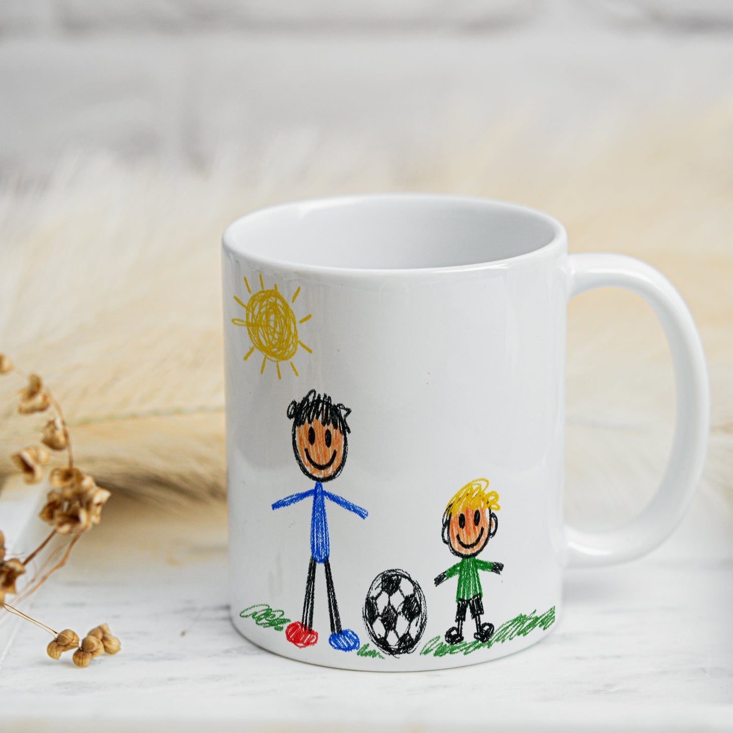 White mug with child-like drawings of two stick figures and a ball on a light wooden surface.