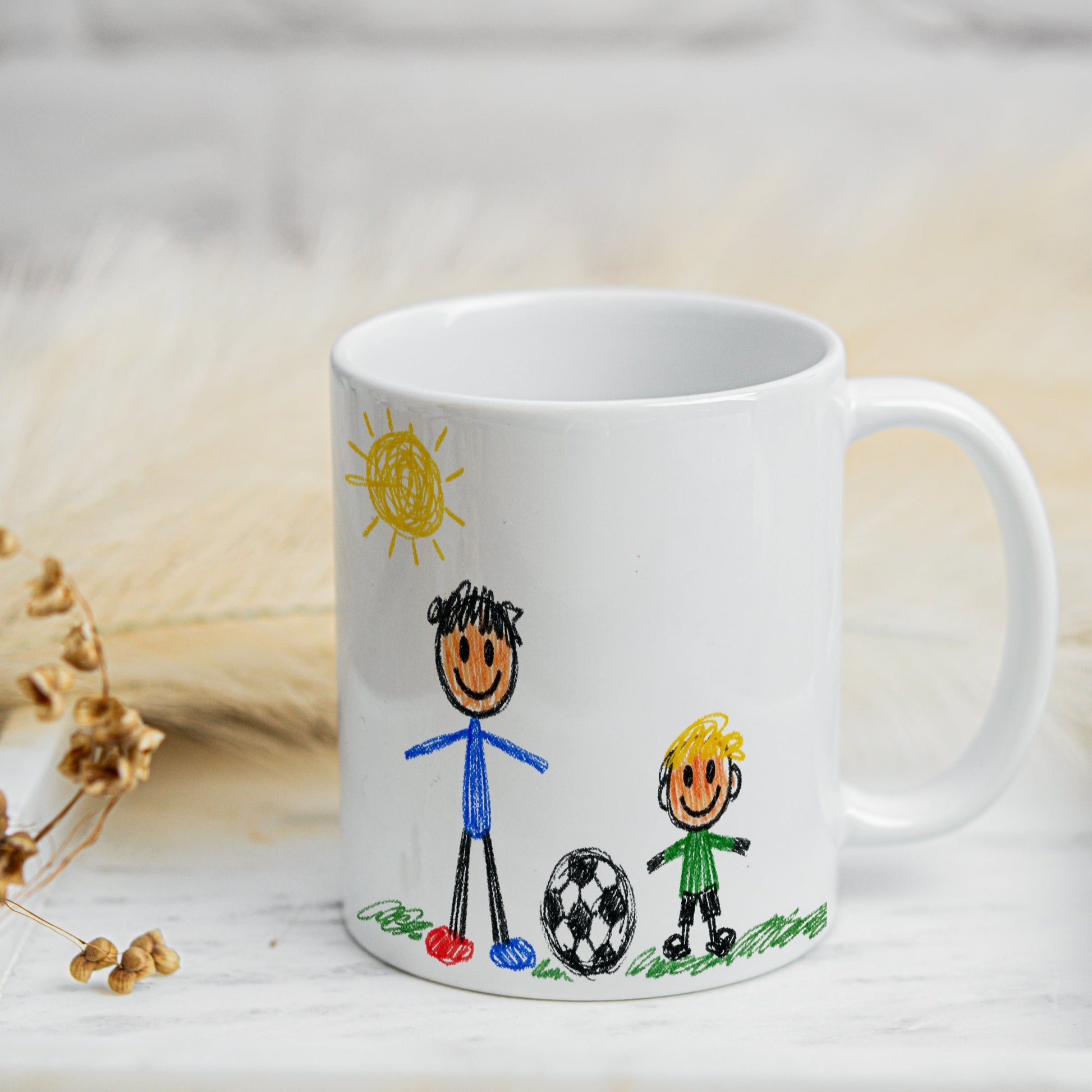 White mug with child-like drawings of two stick figures and a ball on a light wooden surface.
